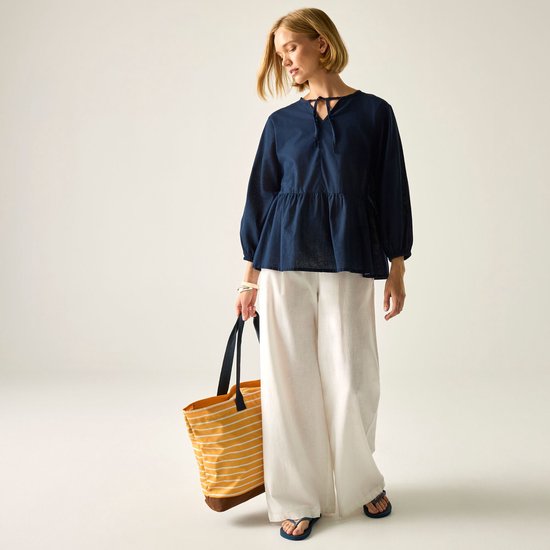 Model draagt een marineblauwe blouse met strikdetail en wijde witte broek met een gestreepte strandtas.