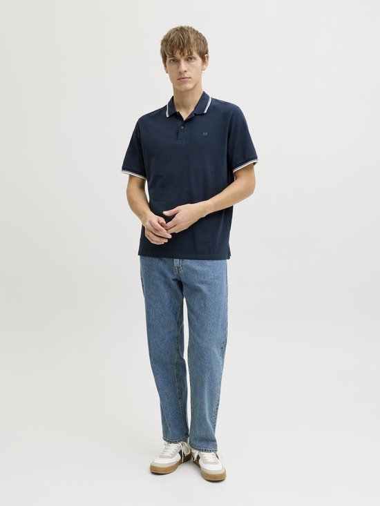 Man in donkerblauw poloshirt met witte streepdetails op kraag en mouwen, en blauwe jeans.