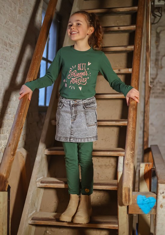 Meisje met bruin paardenstaart, draagt groene longsleeve met tekst, grijze spijkerrok en groene legging.