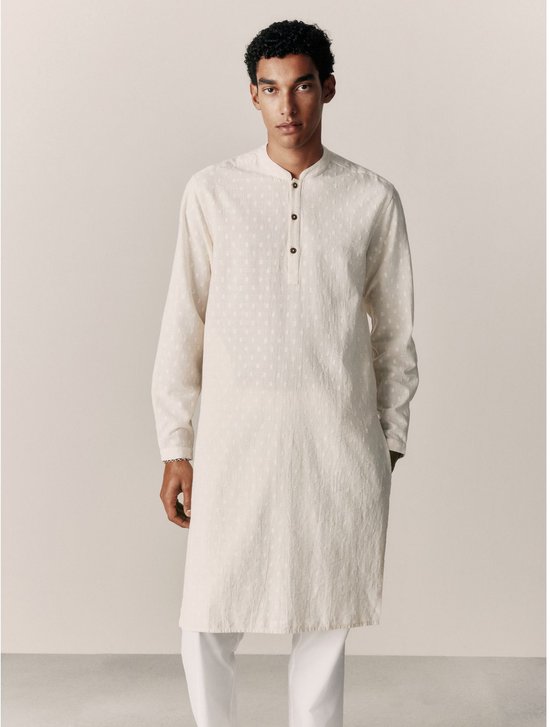 Model draagt een beige kurta-overhemd met dobby-jacquardpatroon, staande kraag en knoopsluiting, gecombineerd met een witte broek.
