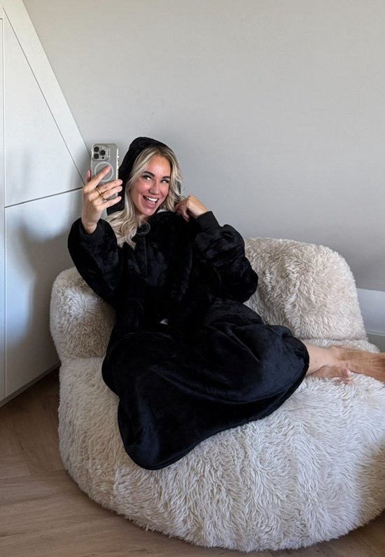 Een lachende vrouw met blonde haren neemt een selfie in een zwarte, oversized hoodie deken, zittend op een grote, fluffy beige poef.