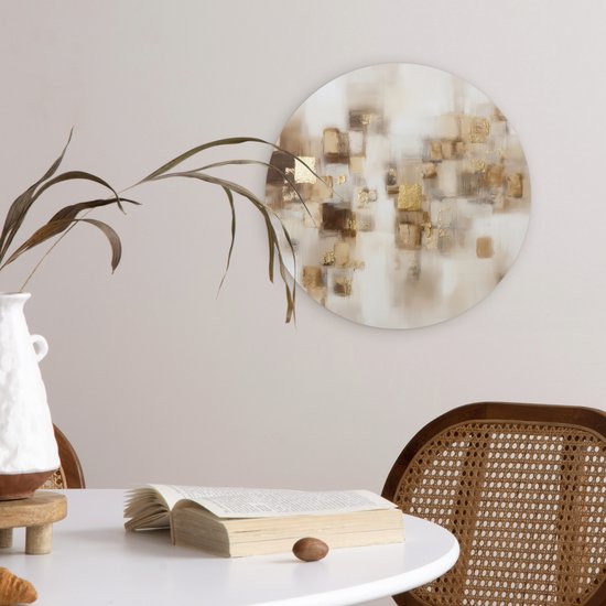 Ronde muurdecoratie met abstracte gouden en bruine vierkanten hangt aan een lichte muur boven een witte tafel.