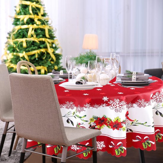 Rond kersttafelkleed met rode ondergrond, witte sneeuwvlokken en een rand met kerstbomen en bloemen. Gedekte tafel bij kerstboom.