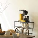 Felgeel Moccamaster koffiezetapparaat met glazen kan gevuld met koffie, op een metalen rek in een keuken.