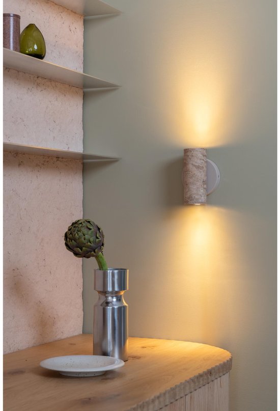Een beige wandlamp van steen met twee lichtbundels op een houten meubel met plateau, vaas met artisjok en een schoteltje.