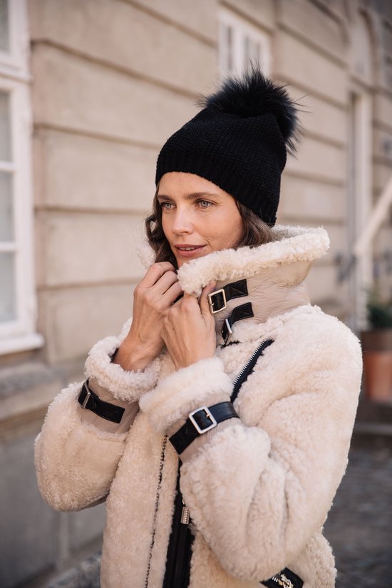 Femme aux longues boucles brunes et bonnet noir à pompon, porte une veste en shearling crème avec des boucles en cuir noir.