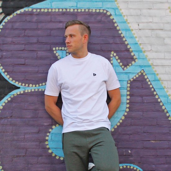 Man met witte T-shirt met korte mouwen en groene joggingbroek, leunend tegen muur met graffiti.