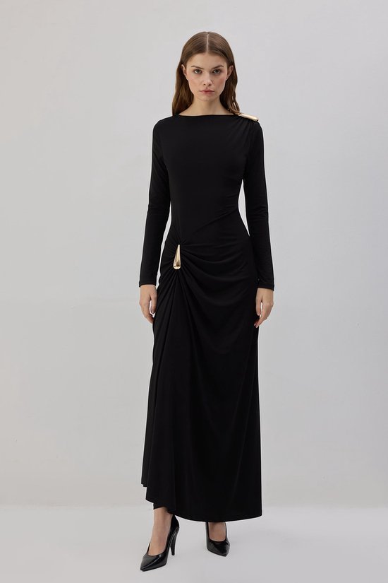 Staand model in een zwarte maxi-jurk met lange mouwen, gouden schouderaccent en gedrapeerde heup met gouden gesp.
