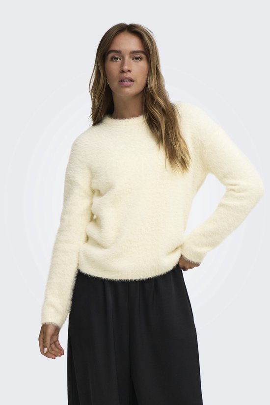 Model in crèmekleurige, pluizige trui met zwarte broek op een witte achtergrond.