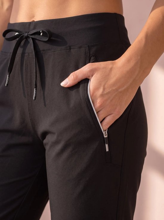Close-up van een hand in een ritszak van een zwarte sportbroek met SJENG merkopdruk op de veters.