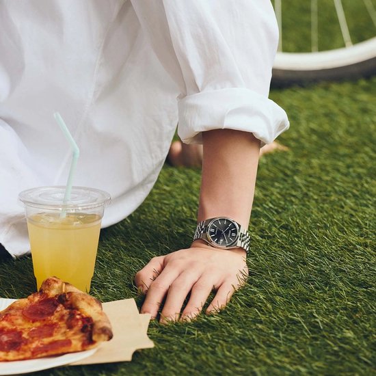 Persoon in wit shirt draagt een zilveren Citizen Tsuyosa horloge met zwarte wijzerplaat in het gras.
