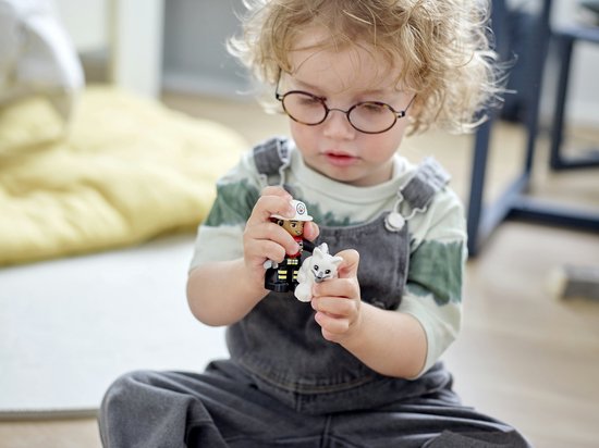 Kind met krullend haar en bril houdt een Duplo brandweerman-figuur en een wit katje vast.