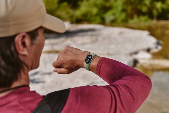Persoon draagt een lichtgroene Fitbit Edge, kijkend naar het scherm bij een stroompje.
