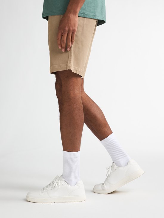 Model draagt beige chino shorts met witte sokken en witte sneakers tegen een witte achtergrond.