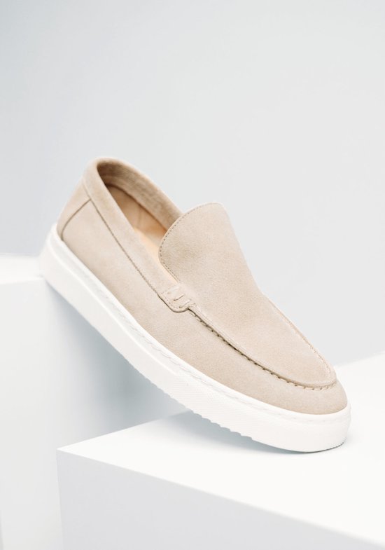 Beige suède loafers met witte zool op een witte ondergrond.