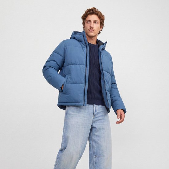 Man met blauwe pufferjas met capuchon, blauwe trui en spijkerbroek, kijkend weg.