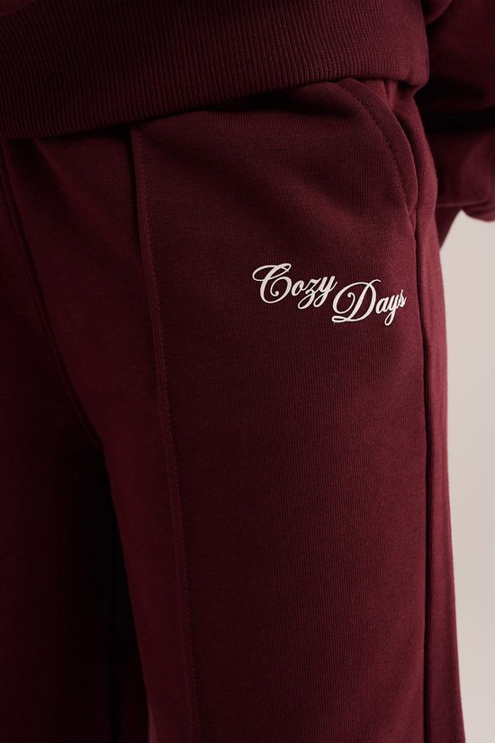 Bordeauxrode joggingbroek met 'Cozy Days' in sierlijk schrift op de zijkant.