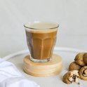 Verre Duralex Picardie avec du café et de la mousse de lait, sur un sous-verre en bois. À côté d'une pile de biscuits.