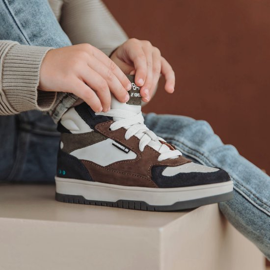 Basket montante marron, blanc et bleu marine, avec lacets. Un enfant attache les lacets.
