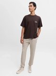 Model draagt een bruin T-shirt met 'Originals Studio' opdruk en beige sweatpants.