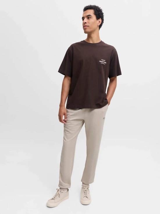Model draagt een bruin T-shirt met 'Originals Studio' opdruk en beige sweatpants.