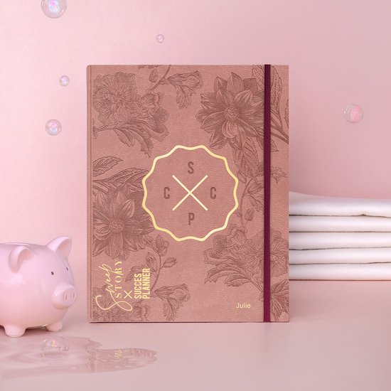 Oudroze agenda met bloemenprint, een gouden cirkel met letters 'S C P' en de naam 'Julie'. Een spaarpot ernaast.