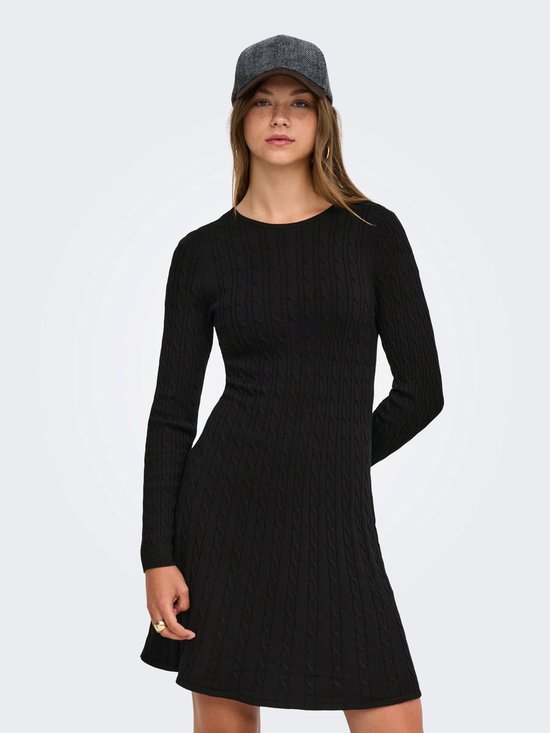 Model met handen achter de rug in een zwarte gebreide mini-jurk met kabels en lange mouwen, met een pet.