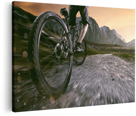 Een wielrenner op een mountainbike rijdt over een onverharde weg met bergen op de achtergrond, bij zonsondergang.