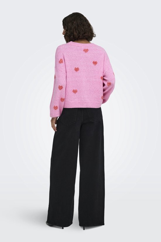 Model draagt achterkant roze trui met rode hartjesprint, lange mouwen en ronde hals, met zwarte broek.