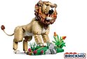 Gele lego leeuw met bruine manen, witte tanden en een groene, stenige omgeving met roze bloemen.
