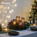 Een adventskrans met vier glazen windlichten met brandende kaarsen, versierd met dennentakken, rode bessen en dennenappels.