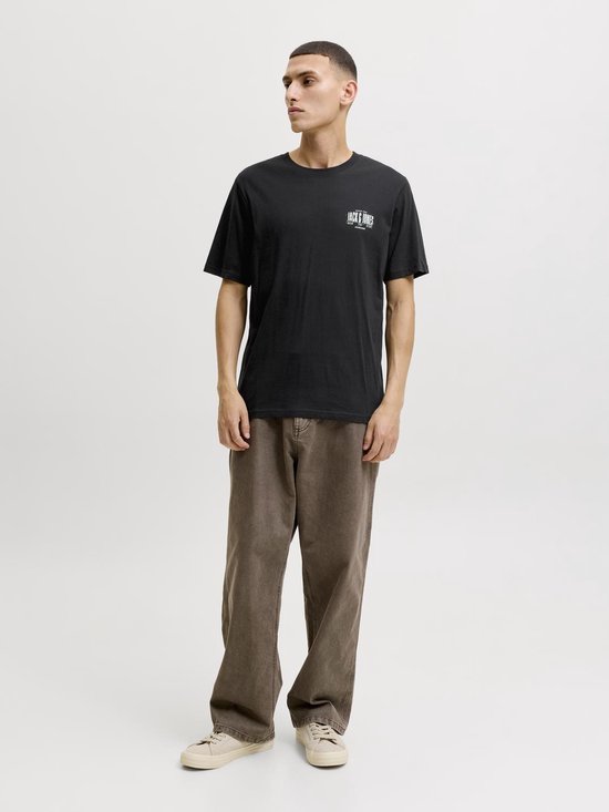 Man draagt een zwarte t-shirt met logo op de borst en bruine wijde broek.