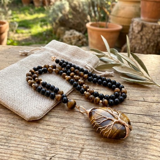 Lange ketting met zwarte en tijgeroogkleurige kralen, met een tijgeroog hanger in boomvorm, naast een linnen zakje op een houten tafel.