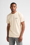 Man staat recht naar voren, draagt een crème T-shirt met opdruk en grijze jeans.