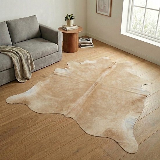 Een grijs-taupe koeienhuid vloerkleed ligt op een houten vloer in een woonkamer met een grijze bank.