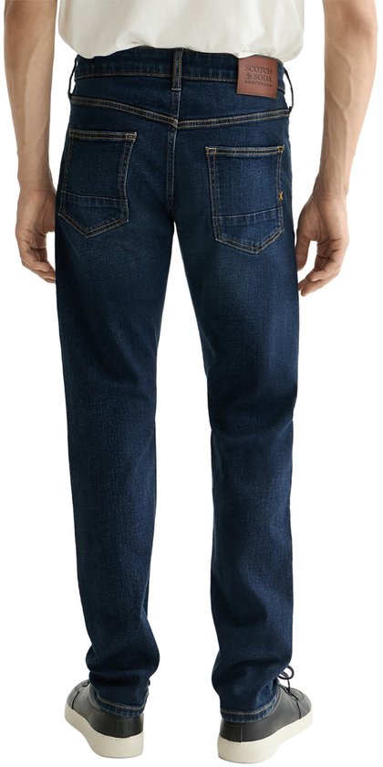 Man met donkerblauwe jeans aan, met gele sierstiksels en bruin label.
