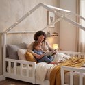 Mère lisant un livre à un enfant dans un lit maison blanc avec barreaux et guirlande lumineuse.