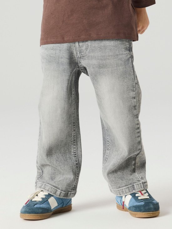 Detail van de benen van een jongen in een grijze spijkerbroek en blauwe sneakers.