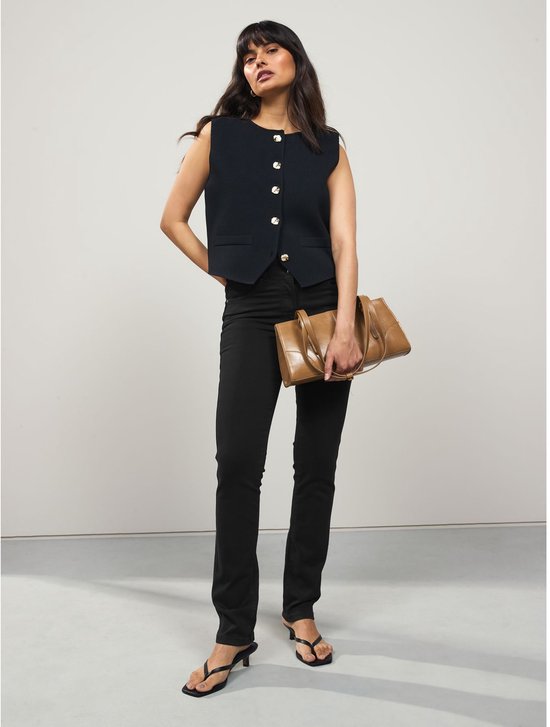 Model draagt een zwarte slim-fit jeans met een donkerblauw vestje en draagt een bruine tas.