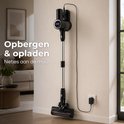 Zwarte steelstofzuiger met zilveren steel hangt aan een muurbevestiging, aangesloten op een lader. "Opbergen & opladen Netjes aan de muur".