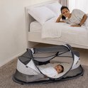 Moeder kijkt vanuit bed naar baby in grijs-wit Deryan campingbedje op de grond.