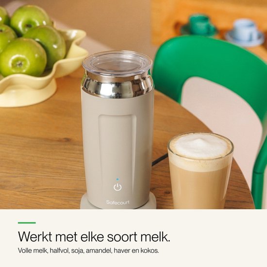 Beige melkopschuimer naast een glas met latte macchiato, op een houten tafel met groene stoel en appels.