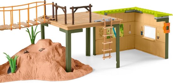 Speelgoed avonturenstation met een grot, hangbrug, huisje op palen met kraan en ladder, en een oranje stoel.