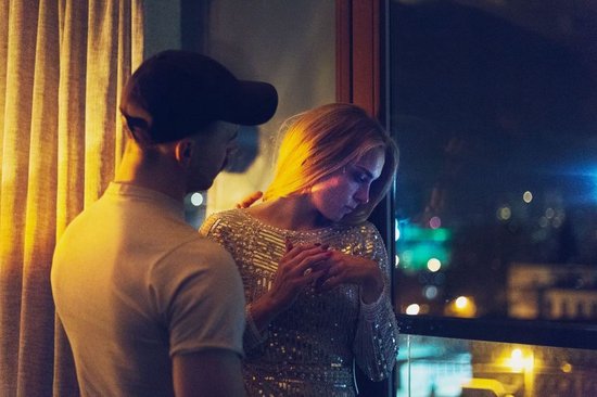 Een man met een pet en een vrouw in een glinsterende jurk staan bij een raam met uitzicht op een stad bij nacht.