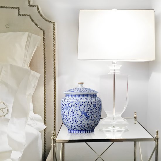 Blauw-witte porseleinen urn met bloemenpatroon op een spiegelende bijzettafel naast een lamp.