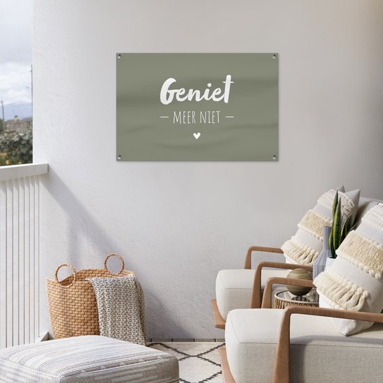 Donkergroene banner met witte tekst 'Geniet - Meer Niet -' en een hartje, hangend aan een witte muur op een balkon met lounge stoelen.