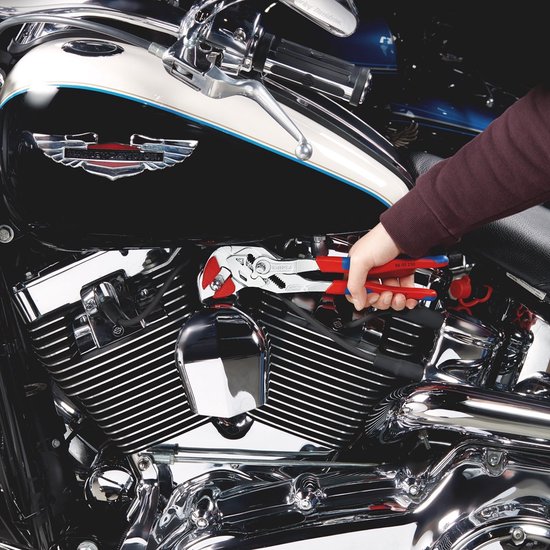 Hand houdt een tang met rode en blauwe handvatten vast, die een deel van een motorfiets aanpast.