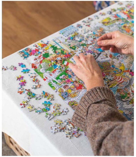 Twee handen leggen de laatste stukjes van een drukke 'Het Tuincentrum' puzzel op een tafel.