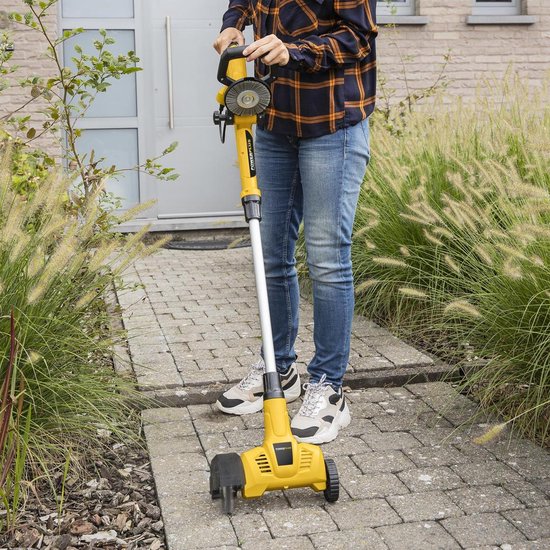 Vrouw gebruikt gele elektrische onkruidverwijderaar op een tegelpad naast groen gras.