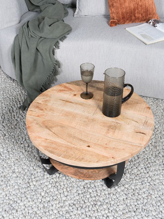 Ronde salontafel van mangohout met een zwart metalen onderstel, naast een grijze bank met groene plaid.